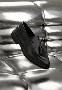 Mustad mustad nahast loafers, millel on narmaste detail, sile tekstuur ja veidi tõstetud tald, paigutatud läikiva hõbedase tausta peale.