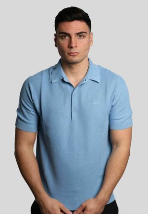 Polo shirt azzurro chiaro a maniche corte con tessuto texturizzato, due bottoni al colletto e polsini a coste, esposto su uno sfondo grigio.