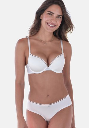 Lächelnde Frau mit langen Haaren, die einen weißen gepolsterten BH und passende Unterwäsche vor einem einfarbigen hellen Hintergrund trägt.