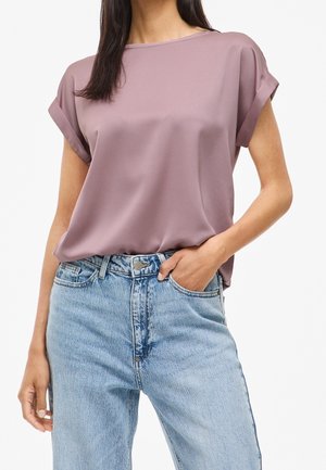 Kobieta ubrana w krótką bluzkę w kolorze mauve, wpuszczoną w wysokowodne jasnoniebieskie dżinsy, z jedną ręką w kieszeni, na tle jednolitego białego tła.