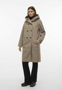 Cappotto beige con cappuccio, dal design doppiopetto, con grandi bottoni e tasche laterali, abbinato a pantaloni neri e stivali robusti marroni.