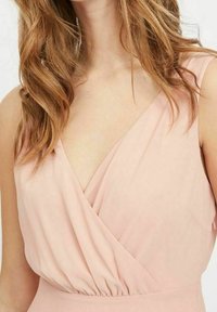 Žena v bezramínkových šatech v jemně růžové barvě s výstřihem do V a nařaseným pasem, s vlnitými světle hnědými vlasy přes ramena.