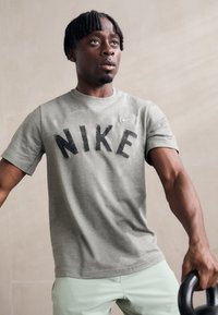 Pelēks kokvilnas T-krekls ar lielu melnu "NIKE" logo priekšpusē. Modelis tur kettlebellu, valkājot gaiši zaļas šortus.