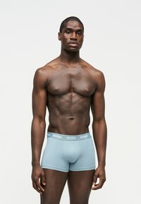 Homme musclé debout, vêtu d'un boxer Hugo bleu clair, sur un fond blanc uni.