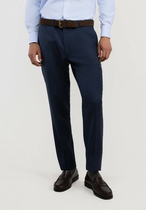 Hombre con pantalones de vestir azul marino, camisa azul clara, cinturón marrón, calcetines oscuros y mocasines de cuero marrón oscuro, de pie frente a un fondo liso.