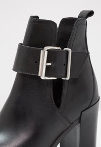 Primer plano de una elegante bota de tobillo negra con tacón y una correa de hebilla plateada, exhibiendo un acabado de cuero suave y un diseño recortado.