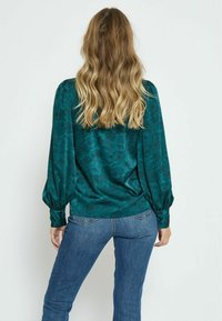 Türkise Bluse mit floralem Muster, langen Puffärmeln und geknöpften Manschetten, kombiniert mit blauen Jeans. Glatte, glänzende Stofftextur.