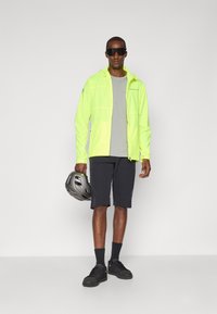 Neon-gelbe, leichte Jacke mit Kapuze, graues T-Shirt, schwarze Shorts, schwarze Socken und Schuhe, hält einen grauen Fahrradhelm.