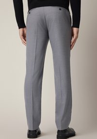 Szare, dopasowane spodnie na miarę o wąskim kroju, z tylnymi kieszeniami zapinanymi na guziki i subtelną teksturą, zestawione z czarnym obuwiem.