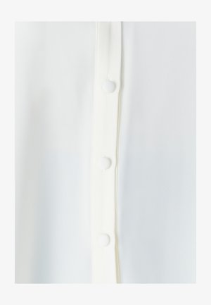 Bijela bluzica s glatkim, blago teksturiranim materijalom, s prednjim gumbima i okruglim bijelim gumbima duž placketa.