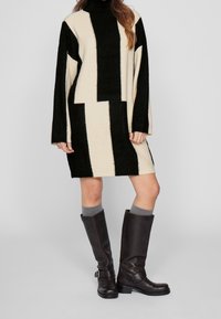 Robe oversized en tricot noir et crème avec motifs géométriques, col montant et manches longues. Bottes marron montantes jusqu'aux genoux et chaussettes grises complètent le look.