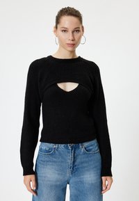 Schwarzer gerippter Strickpullover mit weitem Ausschnitt und Ausschnittdetails auf der Brust. Lange Ärmel. Kombiniert mit hellblauen, weit geschnittenen Jeans.