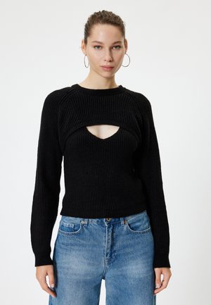 Pull en tricot côtelé noir avec un décolleté large et un détail découpé au niveau de la poitrine. Manches longues. Associe à un jean en denim bleu clair à jambes larges.