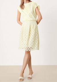 Světle žlutý top s krátkým rukávem, doplněný sukní z krajky po kolena s kulatými výřezy a vlnitým lemem. Nosené s béžovými sandály.