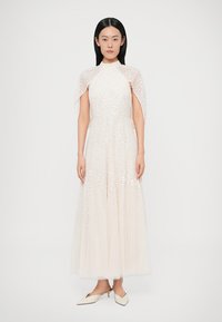 Biele šaty s textúrovaným prekladom, dĺžka po zem, vysoký výstrih a rukávy pripomínajúce plášť. Hladká tkanina s detailmi vzoru, nosené s minimalistickými topánkami.