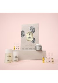 El Set Aniversario 70 Años de Babor incluye botellas y ampollas en un empaque blanco y dorado, junto con un frasco de crema, exhibidos sobre un fondo rosa.