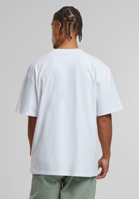White cotton t-shirt with a relaxed fit, short sleeves, and a crew neckline. Visible stitching details on the hem, worn with green trousers.