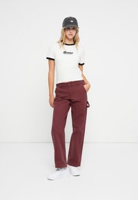 Hvit T-skjorte med svart kant og "Dickies" grafikk, kombinert med burgunderbukser og hvite joggesko, toppet med en mørkegrå caps.