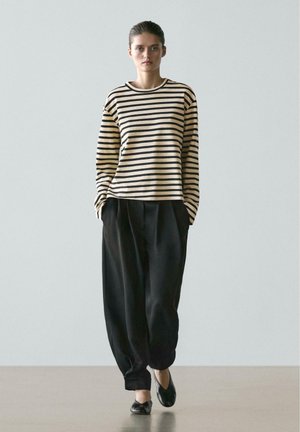 Femme marchant avec assurance, portant un haut à manches longues beige et noir à rayures, un pantalon noir ample et des chaussures plates noires sur un fond neutre.