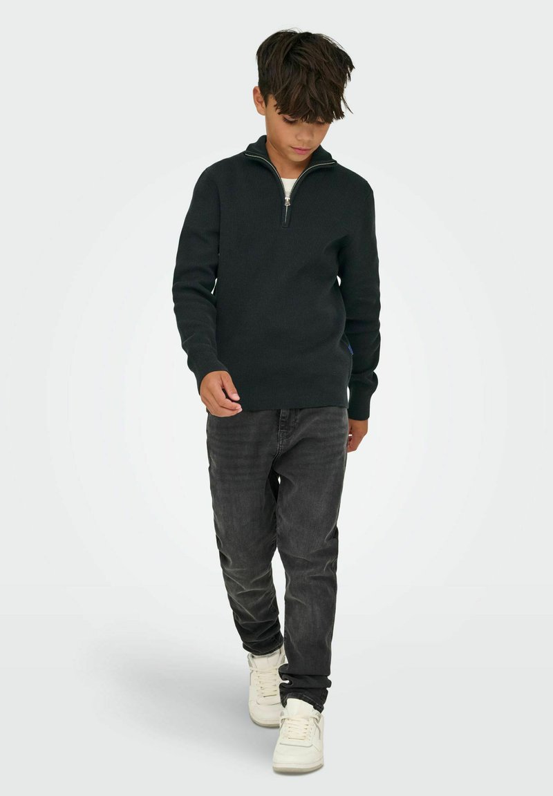 Pull en tricot noir avec un col zippé à un quart, associé à un jean gris et des baskets blanches. Le pull présente des poignets et un ourlet côtelés.