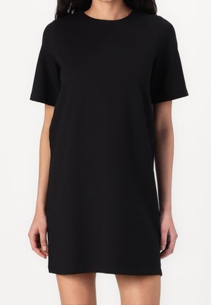 Kobieta w prostej czarnej sukience z krótkim rękawem, sięgającej do kolan, na białym tle, z rozluźnionymi ramionami po bokach.