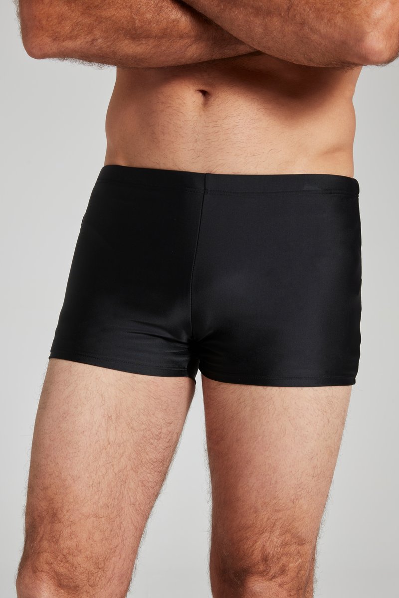 Torace maschile e parte superiore delle gambe che indossano pantaloncini da bagno neri aderenti, braccia incrociate sopra la vita, in piedi contro uno sfondo chiaro e semplice.