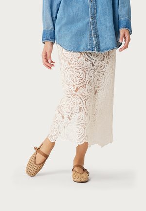 Persona che indossa una camicia di jeans blu, una gonna bianca in pizzo alla caviglia e ballerine Mary Jane color cuoio intrecciate con cinturino e fibbia.