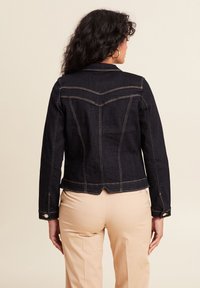 Donkere spijkerjas met een aansluitend ontwerp, contrasterende gele stiksels en een ruggeometrie.detail. Voorzien van knoopsluitingen bij de mouwen en een cropped zoom.