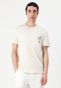 T-shirt beige à manches courtes en coton, avec un petit motif sur la poitrine gauche et un logo sur la manche.