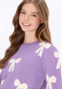 Lila Pullover mit weißen Schleifenmustern, runder Halsausschnitt und weicher Textur, ergänzt durch subtile funkelnde Details.