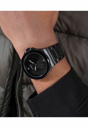 Czarny metalowy zegarek na nadgarstek z minimalistyczną tarczą i okienkiem daty, noszony na lewym nadgarstku na czarnym rękawie i szarej pikowanej kurtce.