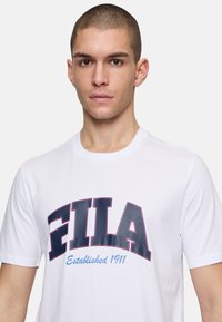 Camiseta de algodón blanca con un prominente logo de "FILA" en azul marino y "Establecido en 1911" en azul claro, cuello redondo y mangas cortas.