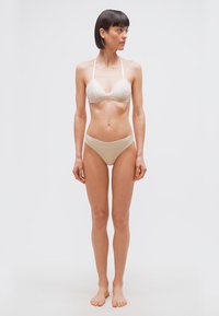 Γυναίκα με κοντά σκούρα μαλλιά που φοράει μπεζ σουτιέν και εσώρουχα, όρθια ξυπόλυτη μπροστά σε ένα απλό γκρι φόντο, κοιτάζοντας προς τα αριστερά της.