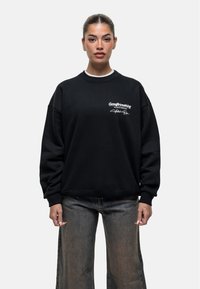 Felpa nera oversize con polsini e colletto a coste. Presenta testo ricamato bianco sul petto. Indossata con jeans chiari a gamba larga.
