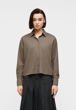 Frau mit glatten dunklen Haaren trägt ein taupefarbenes, langärmliges Hemd mit Knöpfen und einen dunklen, plissierten Rock, steht vor einem schlichten, hellen Hintergrund.