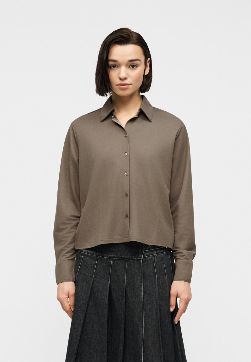 Γυναίκα με ίσια σκούρα μαλλιά φοράει μια ταμπά πουκαμίσα με μακριά μανίκια και κουμπιά και μια σκουρόχρωμη πλισέ φούστα, στέκεται μπροστά σε ένα απλό φωτεινό φόντο.