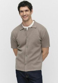 Korte mouwen, beige gebreid knoopshirt met een kraag en grijze knopen, gecombineerd met donkere jeans, op een model tegen een effen achtergrond.
