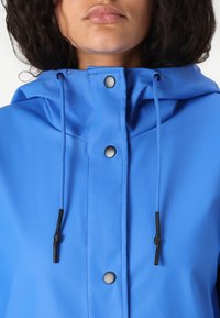 Veste de pluie bleue avec capuche, boutons et cordons noirs. Le matériau a une texture lisse avec une finition mate.