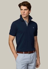 Homme portant un polo bleu marine avec le col relevé et un pantalon rayé blanc et bleu, debout avec une main dans la poche, regardant vers la gauche.