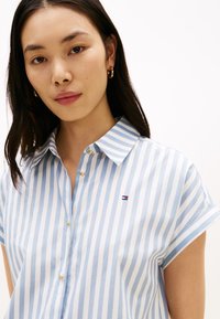 Ανοιχτό μπλε και λευκό ριγέ κοντομάνικο πουκάμισο με μυτερό γιακά, που διαθέτει χρυσά κουμπιά και μικρό λογότυπο στο στήθος.