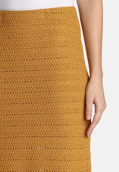 Close-up van een hand die rust naast een mosterdgele, gestructureerde gebreide rok en een wit topje tegen een effen achtergrond.