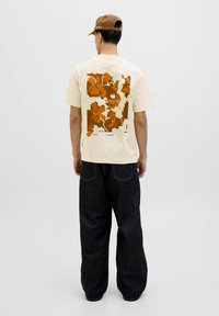 Cream t-shirt with a large orange floral graphic on the back, paired with dark blue wide-leg jeans. Brown cap completes the look.