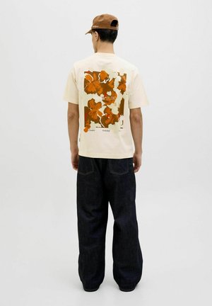 Camiseta crema con un gran gráfico floral naranja en la parte posterior, combinada con jeans de pierna ancha de color azul oscuro. Una gorra marrón completa el look.