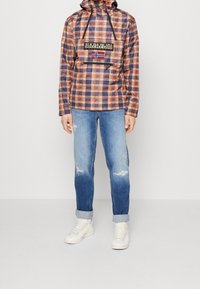 Person iført en orange og lilla ternet hættetrøje med en frontlomme og blå jeans med opsmøg og hvide sneakers, stående på et hvidt gulv.