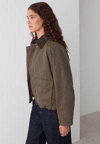Olijfgroene jas van duurzaam materiaal met een bruine corduroy kraag, rits closure en een voorzak. Heeft een relaxte pasvorm en zichtbare stiksels.