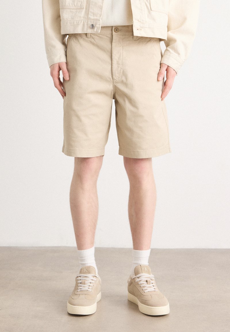 Beige bavlněné šortky s rovný střih, zapínáním na knoflík a bočními kapsami, doplněné o béžové tenisky a bílé kotníkové ponožky.