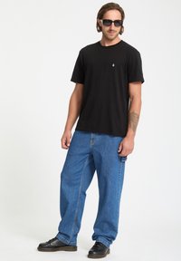 Camiseta de algodón negra con mangas cortas y un pequeño logo blanco, combinada con jeans de mezclilla azules y holgados, y zapatos negros.
