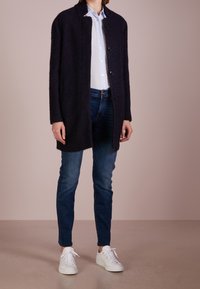 Abrigo largo de textura oscura con corte recto, con botones negros, llevado sobre una camisa azul clara y jeans oscuros. Combinado con zapatillas blancas.