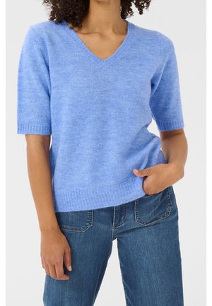 Woman wearing a light blue short-sleeve V-neck sweater and blue jeans, one hand in pocket against a plain background.