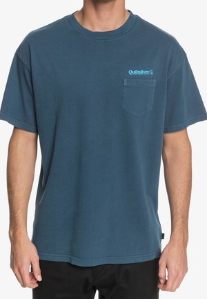 Mand iført blå Quiksilver T-shirt med brystlomme og logo over lommen, kombineret med sorte bukser mod hvid baggrund.